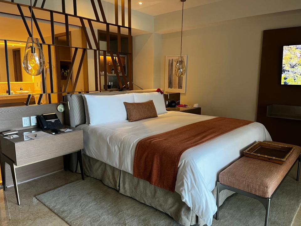 A hotel suite with a wall made of abstract metal poles divides the bathroom from the bedroom. The bed has white shits and a tan blanket on top and is next to a faux wood desk with a phone