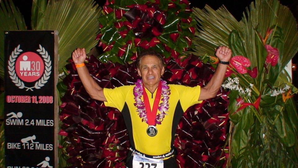 Dr. Joseph Maroon holding his hands in the air as he stands next to an ironman triathlon sign. There is a medal around his neck.