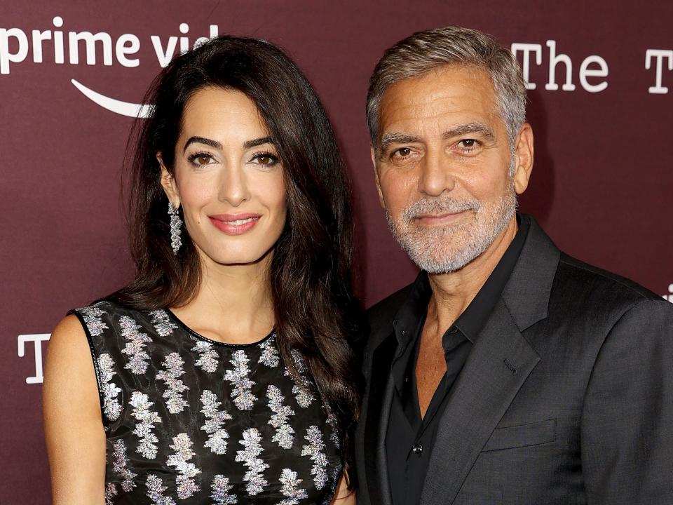Amal Clooney wears a black dress with a silver pattern and George Clooney wears a black suit. Both stand in front of a burgundy background with "Prime Video" text on it