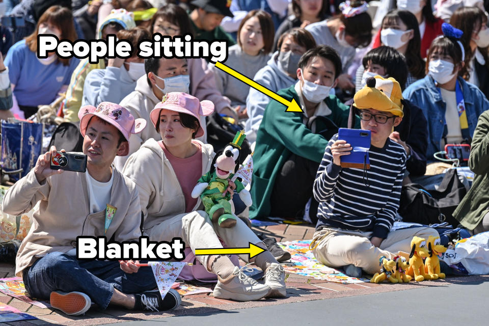 Group of people, some wearing hats and holding Disney-themed items, seated on the ground in a crowded outdoor area