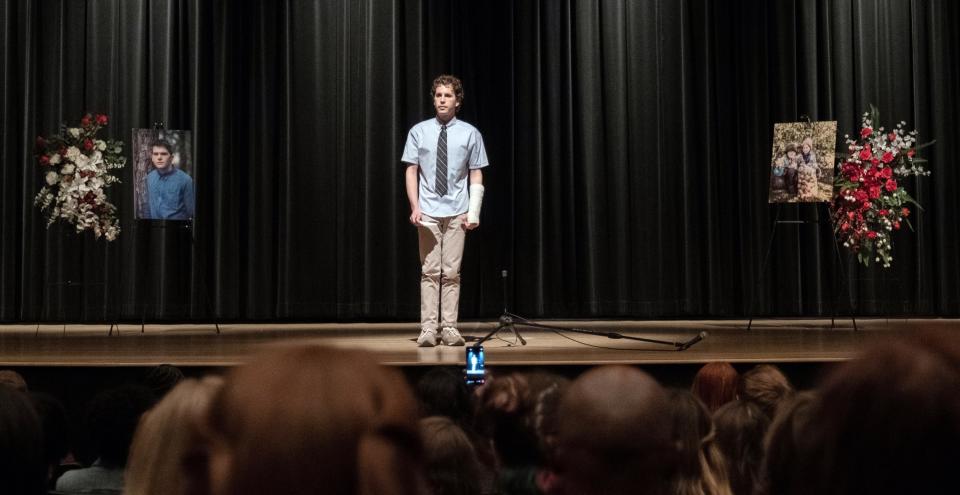 Evan Hansen standing at the mic in a school auditorium with pictures of a fellow student who has died off to the sides