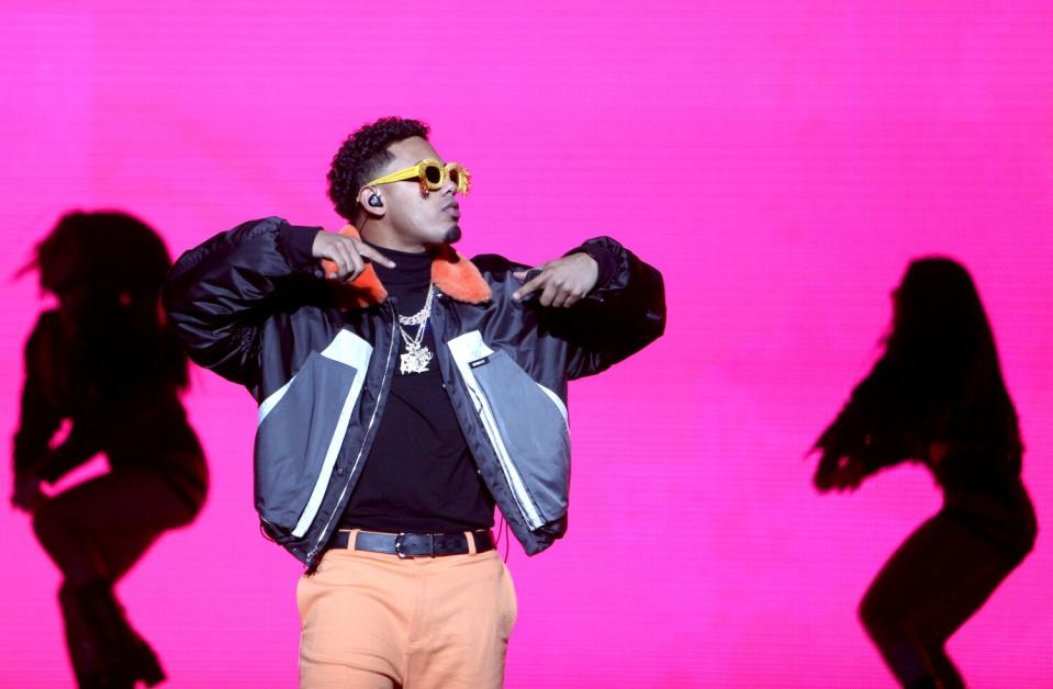 A reggaeton artist performs onstage with a pink background and two backup dancers