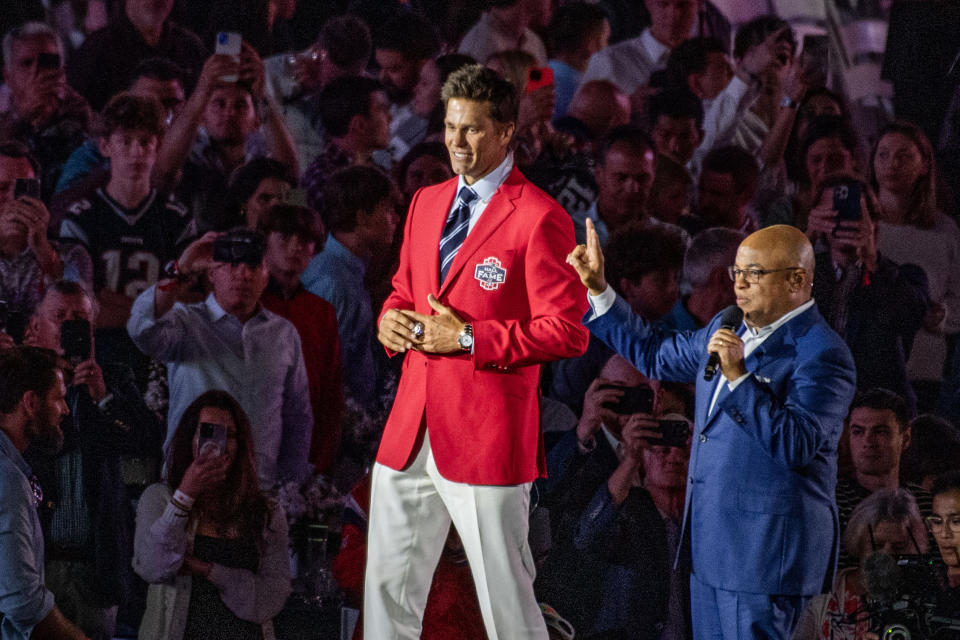 Former New England Patriots quarterback Tom Brady (L) wears his red Patriots Hall of Fame induction jacket as he greets fans, flanked by US sports commentator Mike Tirico, during the 2024 induction ceremony at the Gillette Stadium in Foxborough, Massachusetts on June 12, 2024. Tom Brady is the 35th person to be inducted into the Patriots Hall of Fame.  (Photo by Joseph Prezioso/AFP) (Photo by JOSEPH PREZIOSO/AFP via Getty Images)