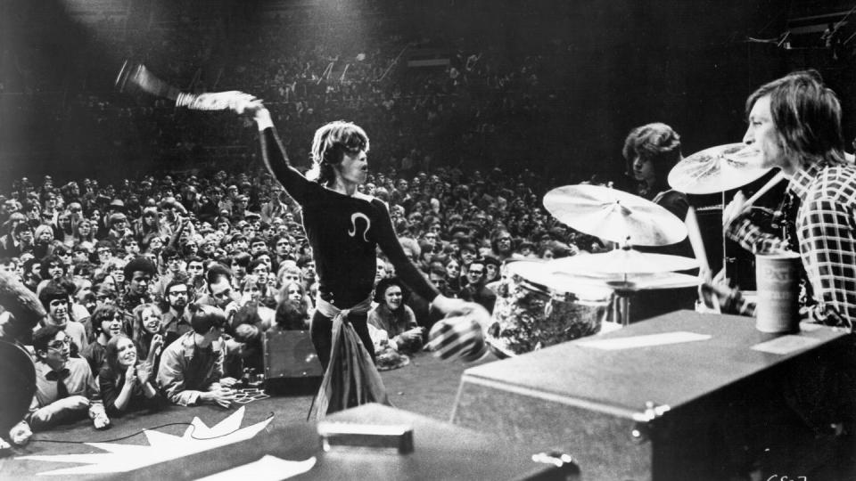 mick jagger whips something around his head while standing on a stage with drumsets and two other men, a crowd of people stands in the background