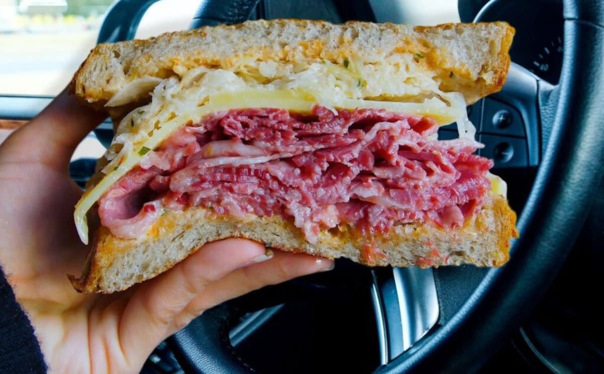 Hand holding Reuben Sandwich, Zingerman's, Ann Arbor, Michigan, steer wheel and window in the background