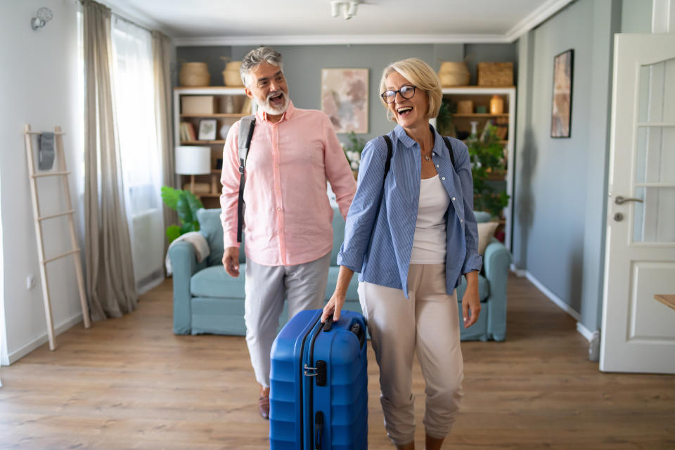 A smiling man and woman in casual clothing carry luggage in a living room, suggesting travel or moving plans