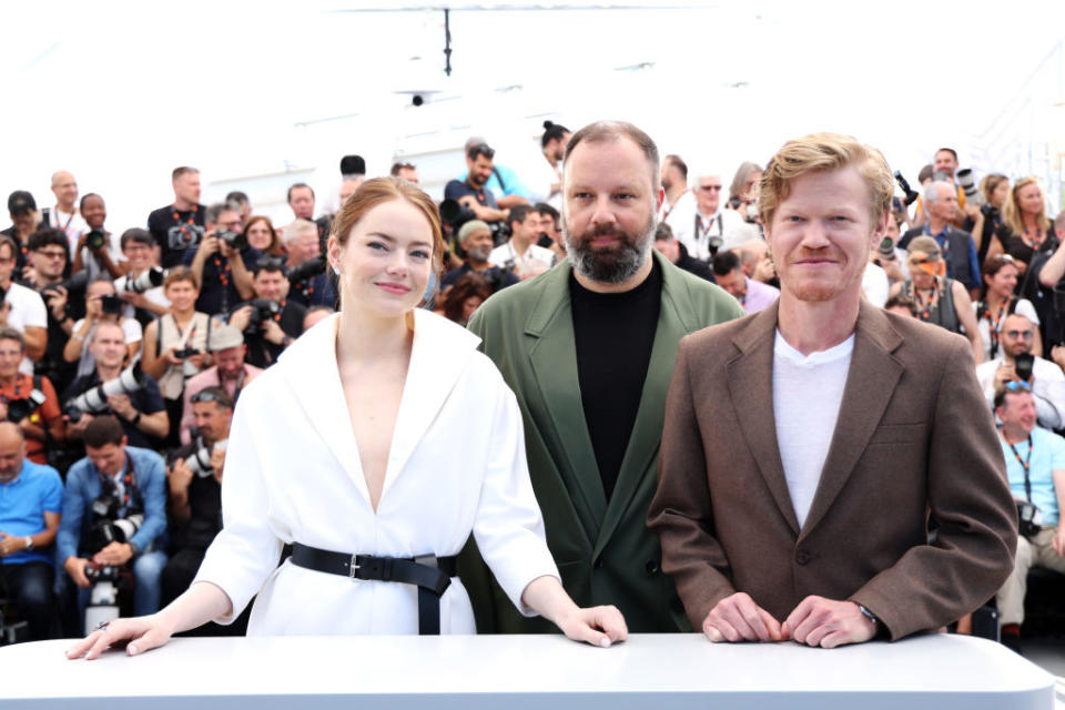 Emma Stone, Yorgos Lanthimos, and Jesse Plemons pose together at a media event, standing behind a table with a crowd of spectators and photographers in the background