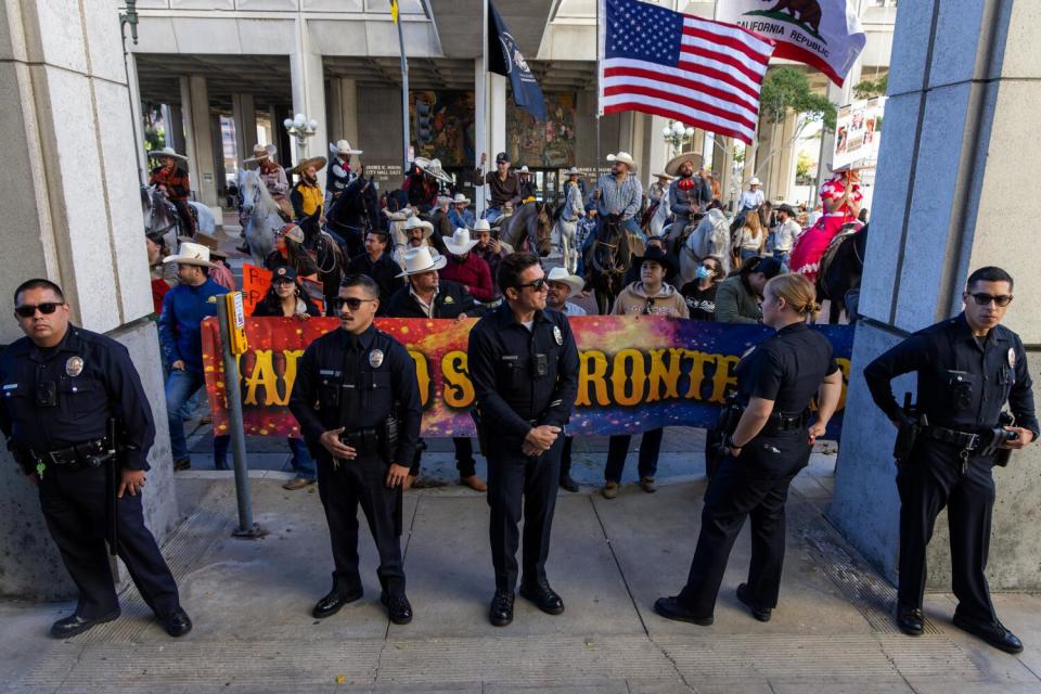 Police officers stand with the backs to a big banner and a crowd behind it.