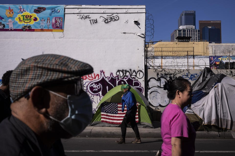 Pedestrians pass by a homeless tent adorned with an American flag across the street from the Los Angeles Mission in the Skid Row area of downtown Los Angeles, Wednesday, Nov. 22, 2023. Downtown's Skid Row neighborhood remains the epicenter of the crisis, where tents extend for blocks and unconscious people can be found splayed on sidewalks and slumped in doorways. Some streets have been transformed into parking lots for rusted, parked RVs. (AP Photo/Jae C. Hong)