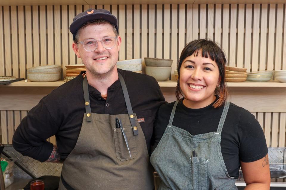 Husband-and-wife chefs stand smiling in a kitchen, wearing aprons