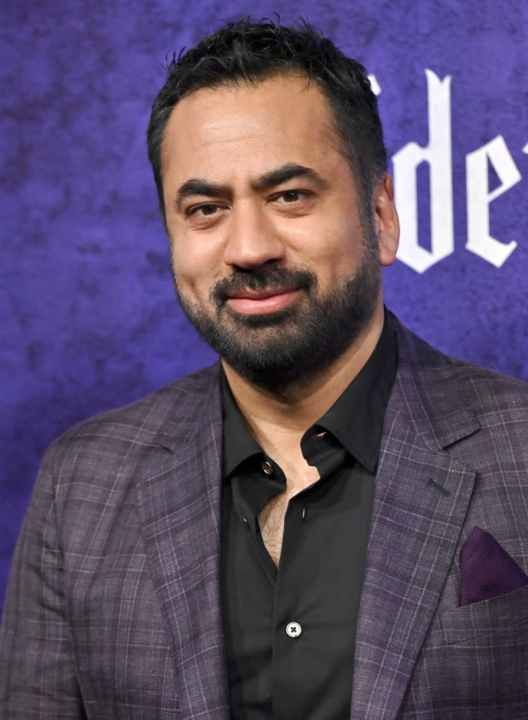 Kal Penn in a checkered suit jacket and black shirt, poses at an event with a purple background