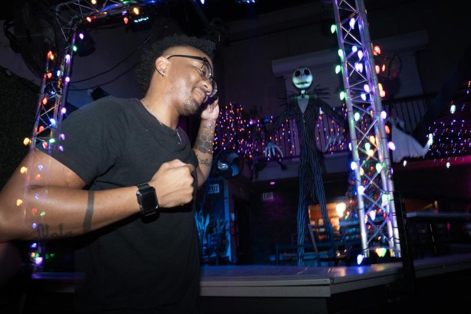 A man in a black T-shirt and glasses stands beneath lights.