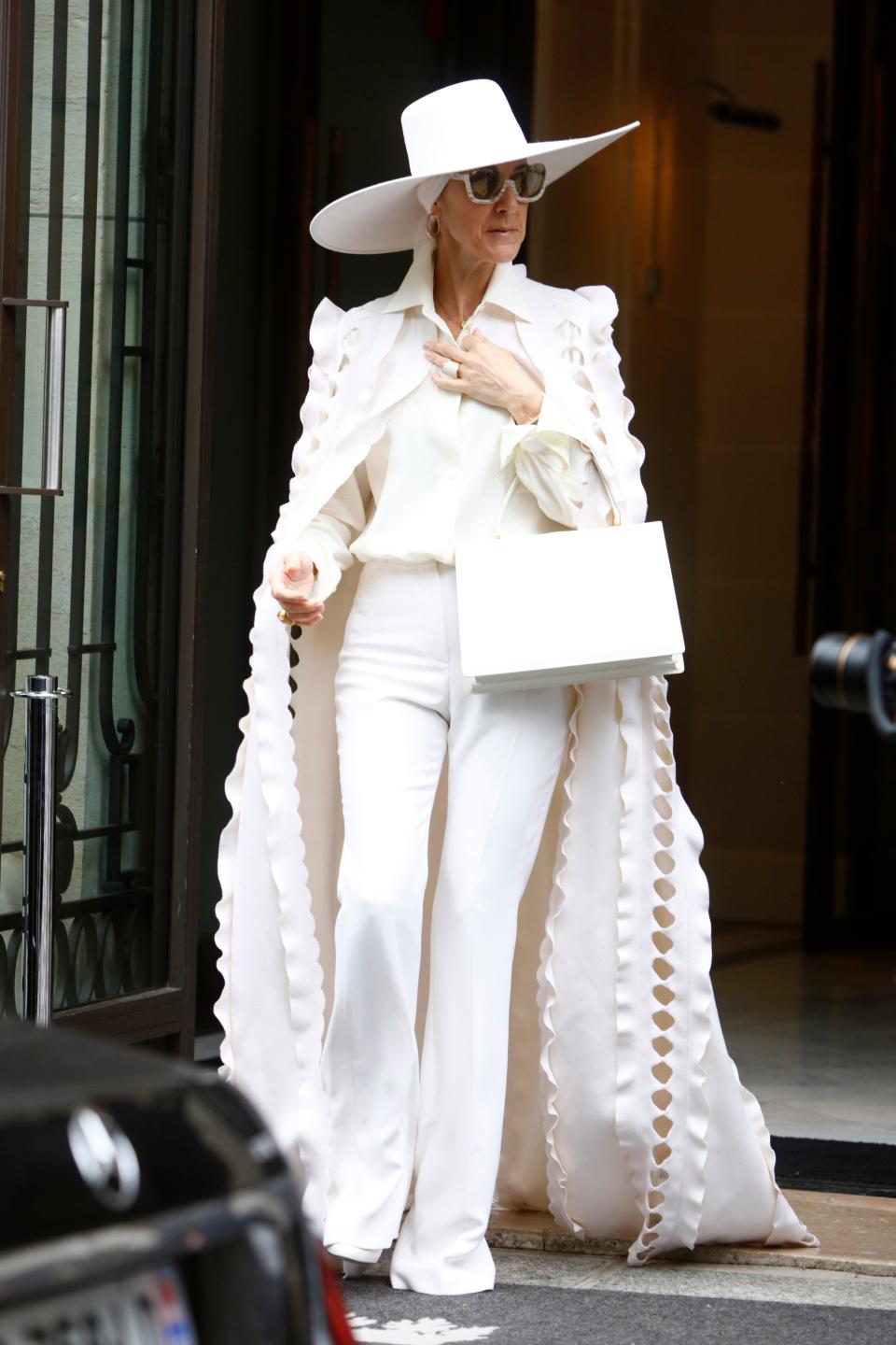 Celine in an all-white look. She's wearing a blouse, high-waisted trousers, a cutout cape, a wide brimmed hat, sunglasses, and a purse.