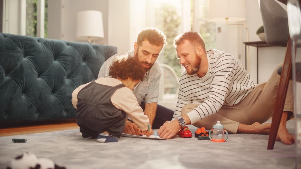 Two men sit on the floor with a small toddler, all playing together as a family
