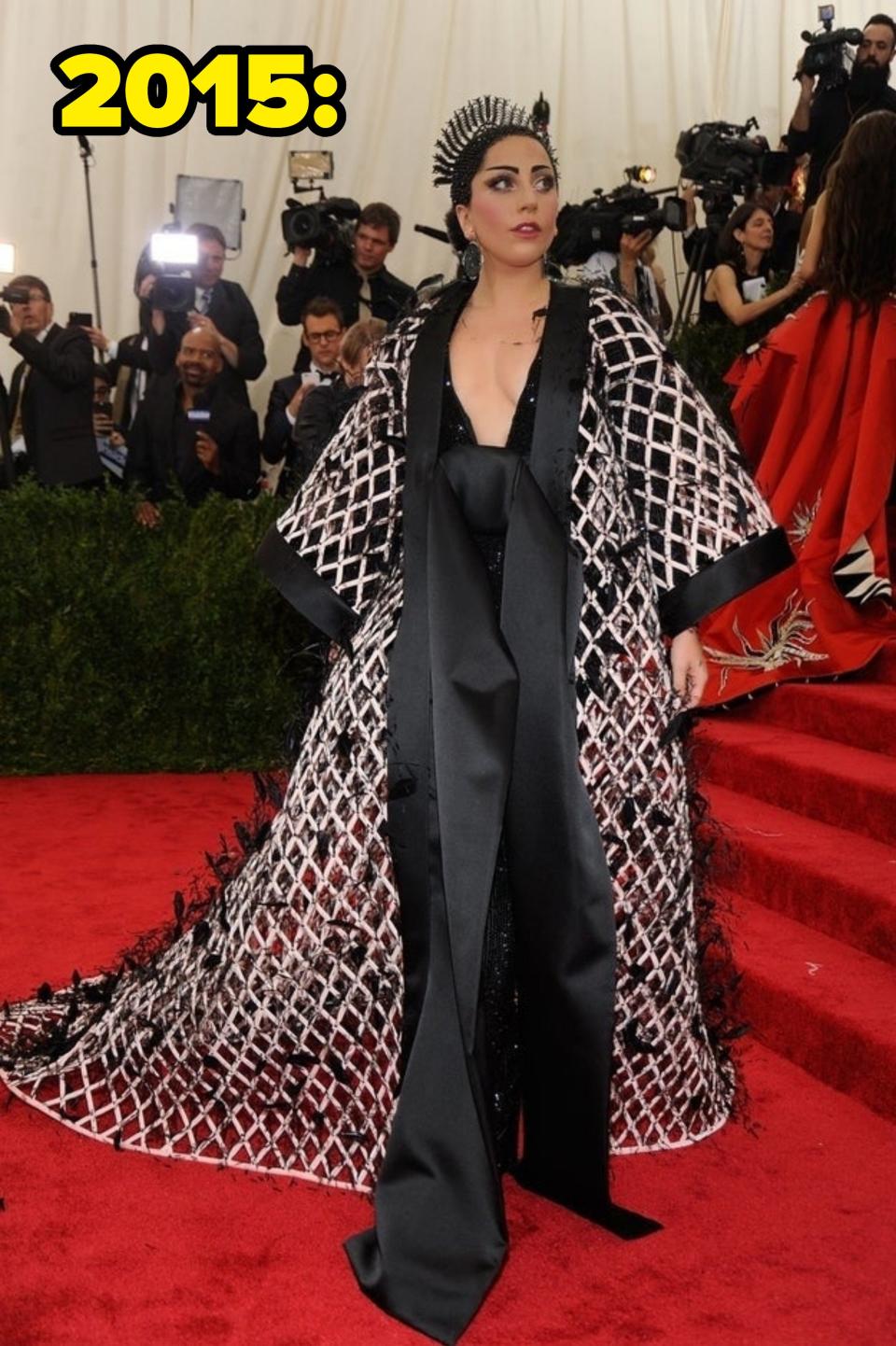 Lady Gaga on the red carpet in an elaborate, patterned gown with a plunging neckline and dramatic cape, wearing a spiked headpiece, surrounded by photographers