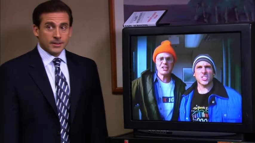 Michael standing next to a TV playing a video of him and Dwight in "The Office"