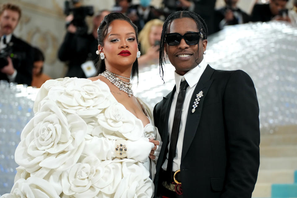 Rihanna, wearing a white gown with 3D roses around her arms and shoulders, poses at the Met Gala with A$AP Rocky, wearing sunglasses and a black suit and tie