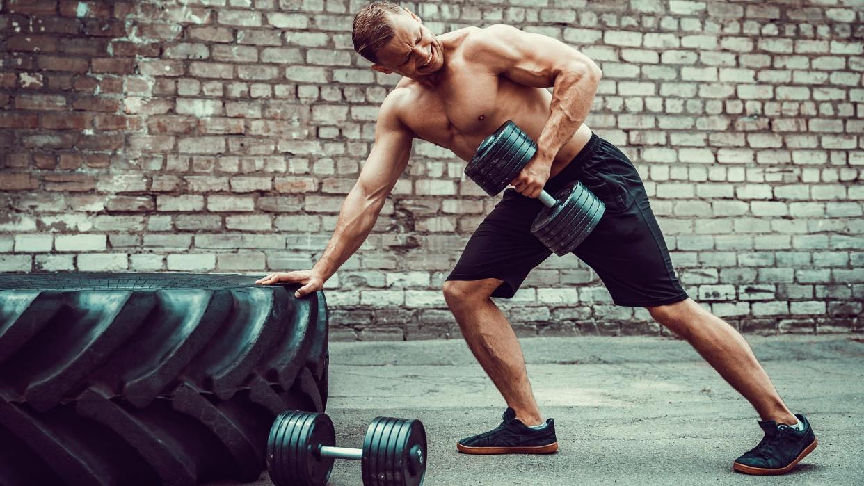  Man leaning against a tyre with one hand and performing a dumbbell bentover row with his left arm. 