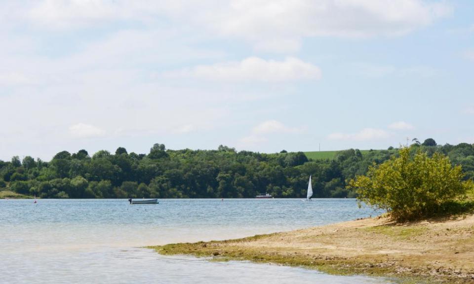 Water’s edge of Bewl Water reservoir in Tunbridge Wells, Kent. An empty boat and a windsurfer are on the lake with a small ferry.