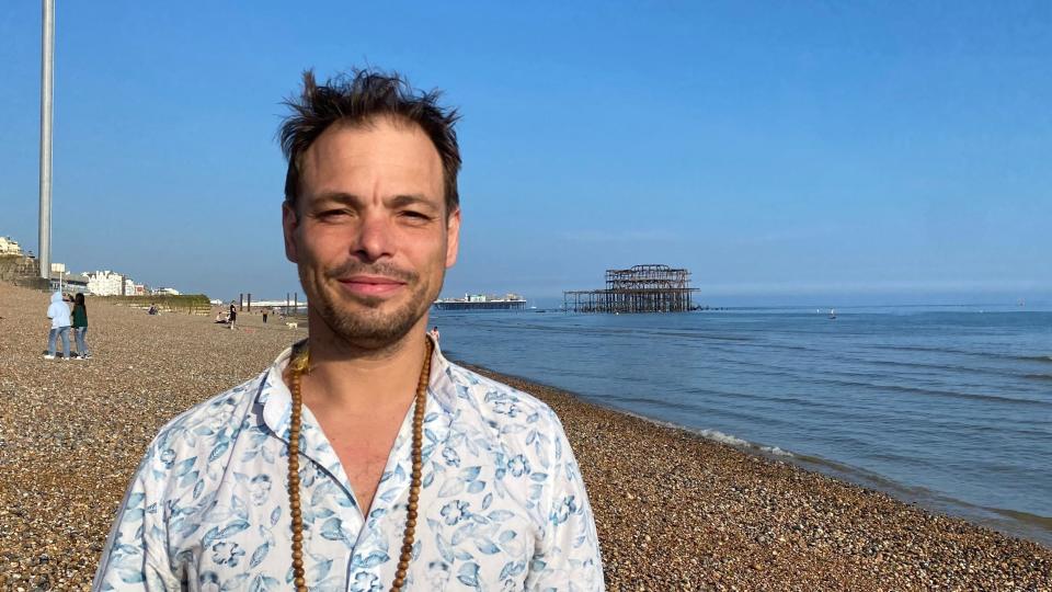 A man wearing a floral shirt with a brown necklace stood on Brighton beach with the West Pier in the background