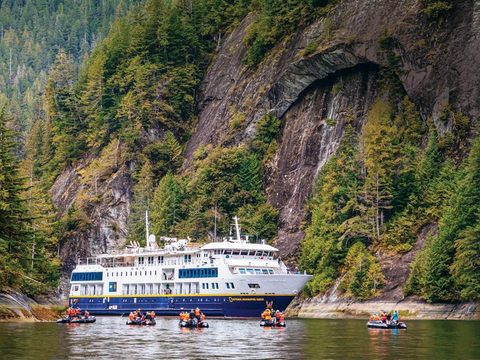 A cruise ship sailing through lush green cliffs.There are people on small boats in front of the cruise ship.