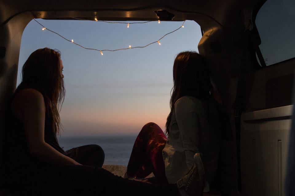 Two people sitting in a van's open trunk, facing a sunset, with a string of lights above