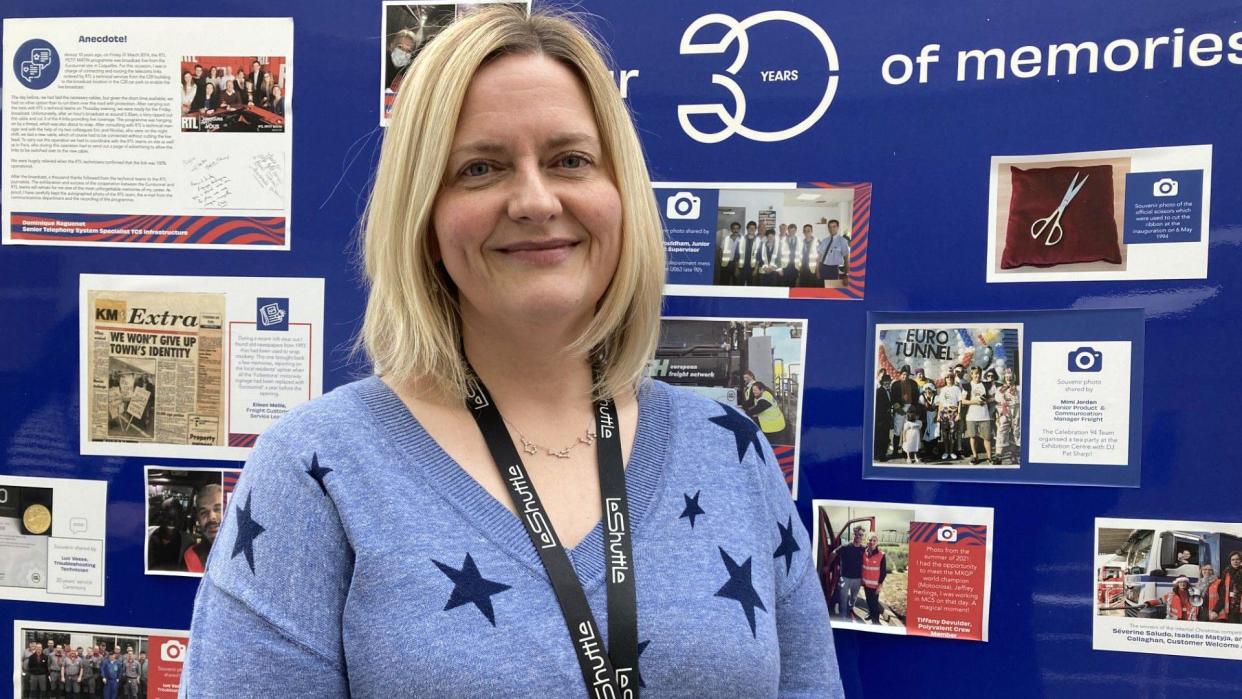 A middle-aged woman with shoulder length blonde hair wears a blue jumper with stars on it and smiles at the camera in front of a board of archive photographs of the Channel Tunnel 