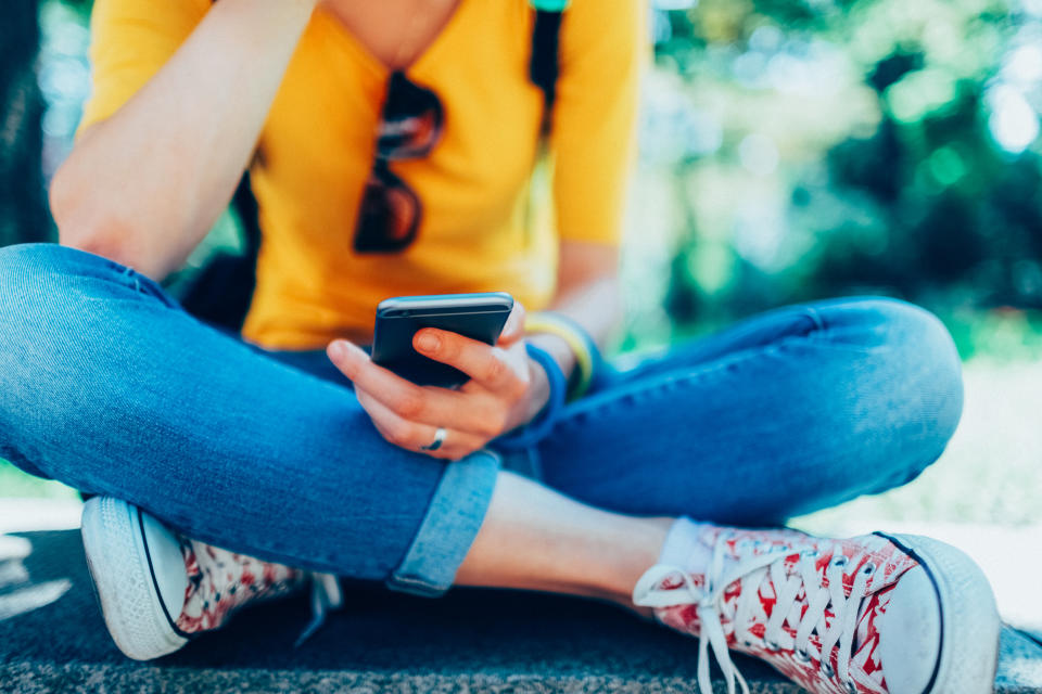 A person is sitting cross-legged outdoors, holding a smartphone and looking at the screen