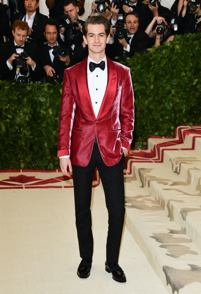 A person in a red velvet jacket and black trousers stands on a carpeted stairway with photographers in the background