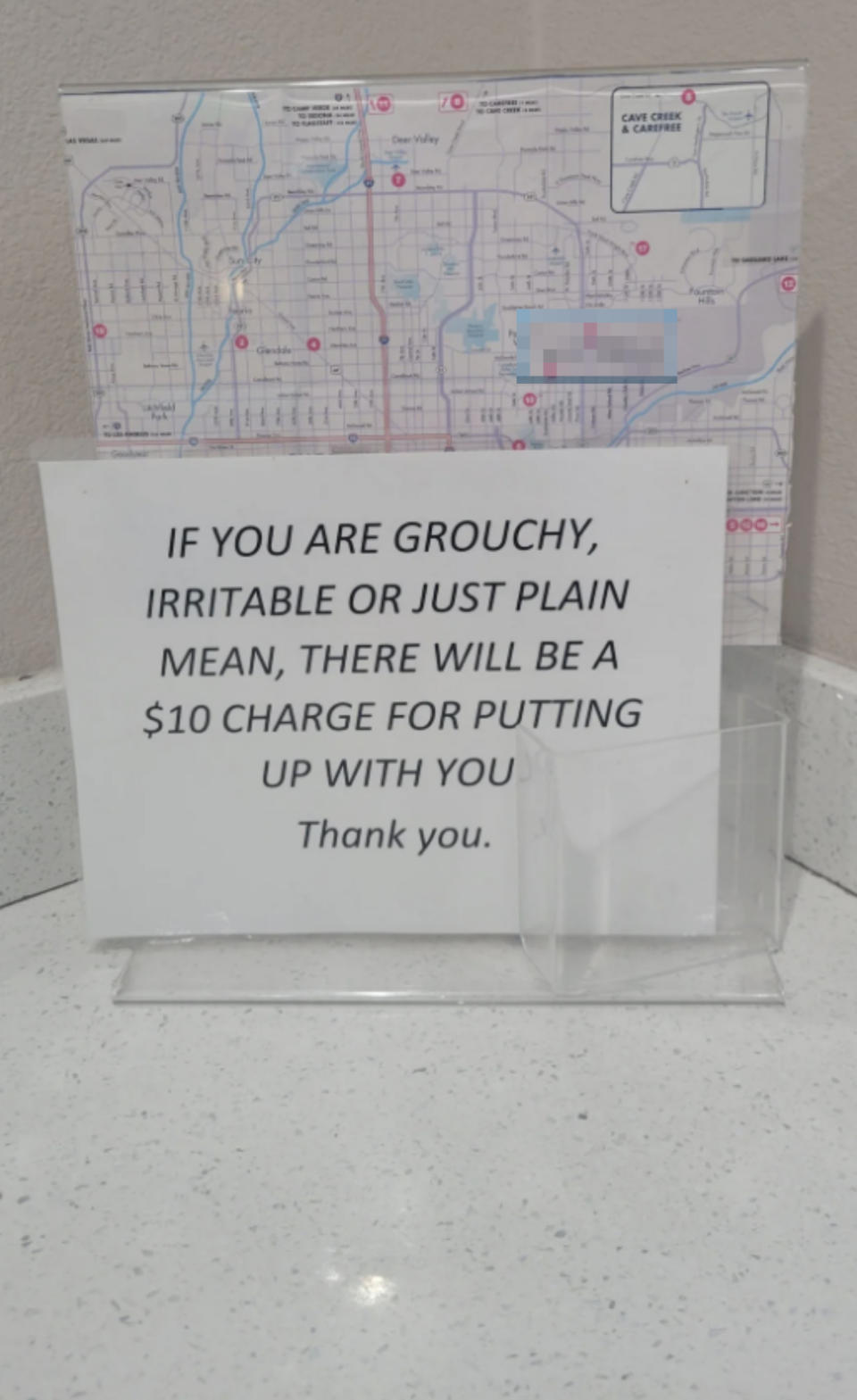 Sign reads: "If you are grouchy, irritable or just plain mean, there will be a $10 charge for putting up with you. Thank you." Background shows a map of Scottsdale