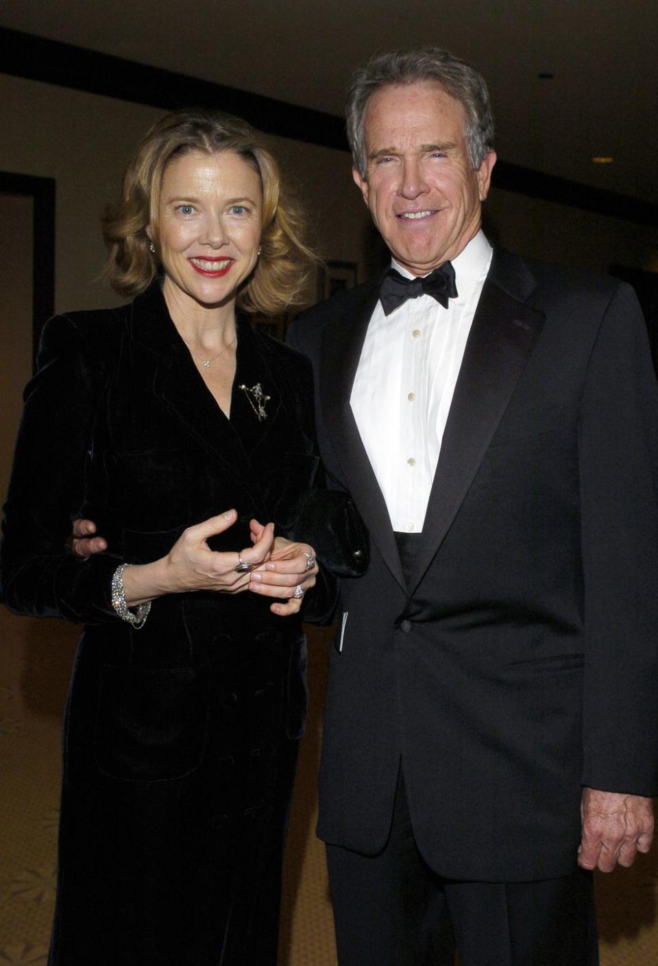 annette bening and warren beatty stand together and smile at the camera, she wears a black long sleeve dress with jewelry, he wears a black tuxedo with a white collared shirt