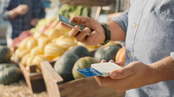 A man at a farmer's market holds a smartphone in one hand and a Square card reader, with credit card inserted, in another.