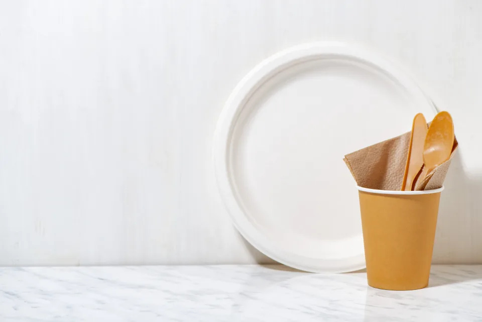 Disposable plate, cup, wooden fork and spoon with a napkin on a white marble surface, typically used in workplaces for eco-friendly meetings or events