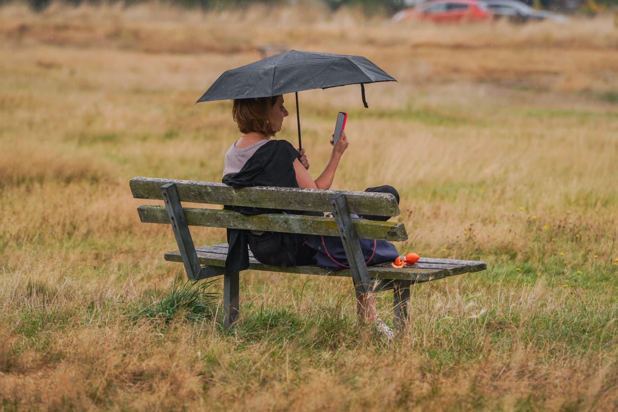 London. 2 September  2024.  A woman sitting on a bench holding a mobile phone with an umbrella during a rain shower on Wimbledon Common, south west London. The forecast is for showery rain and thunderstorms after the warm weather spell which  will affect parts of the UK today. Credit: Amer Ghazzal/Alamy Live News