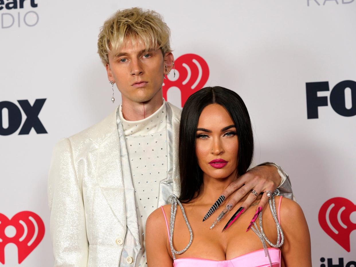 Machine Gun Kelly with his left arm around Megan Fox's shoulder at the red carpet of the iHeartRadio Music Awards.2021