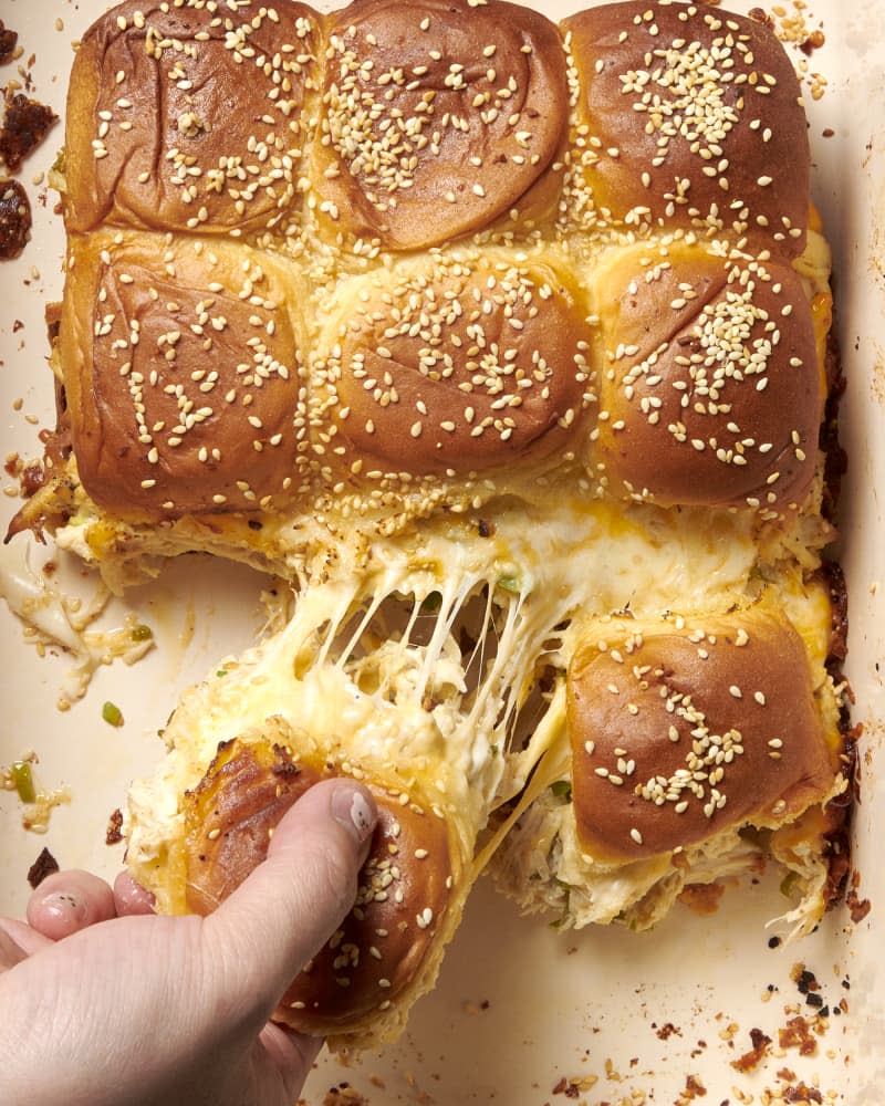 overhead shot of chicken sliders in a baking sheet; a hand pulls one away from chicken slider group showing cheese pull