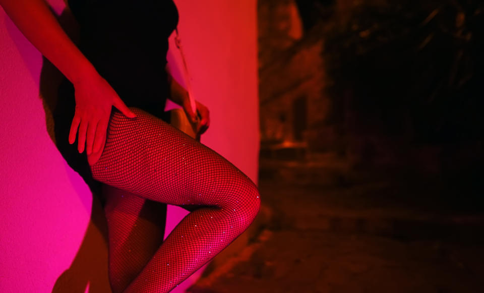 Person in a black outfit and fishnet stockings leaning against a wall in a dimly lit setting