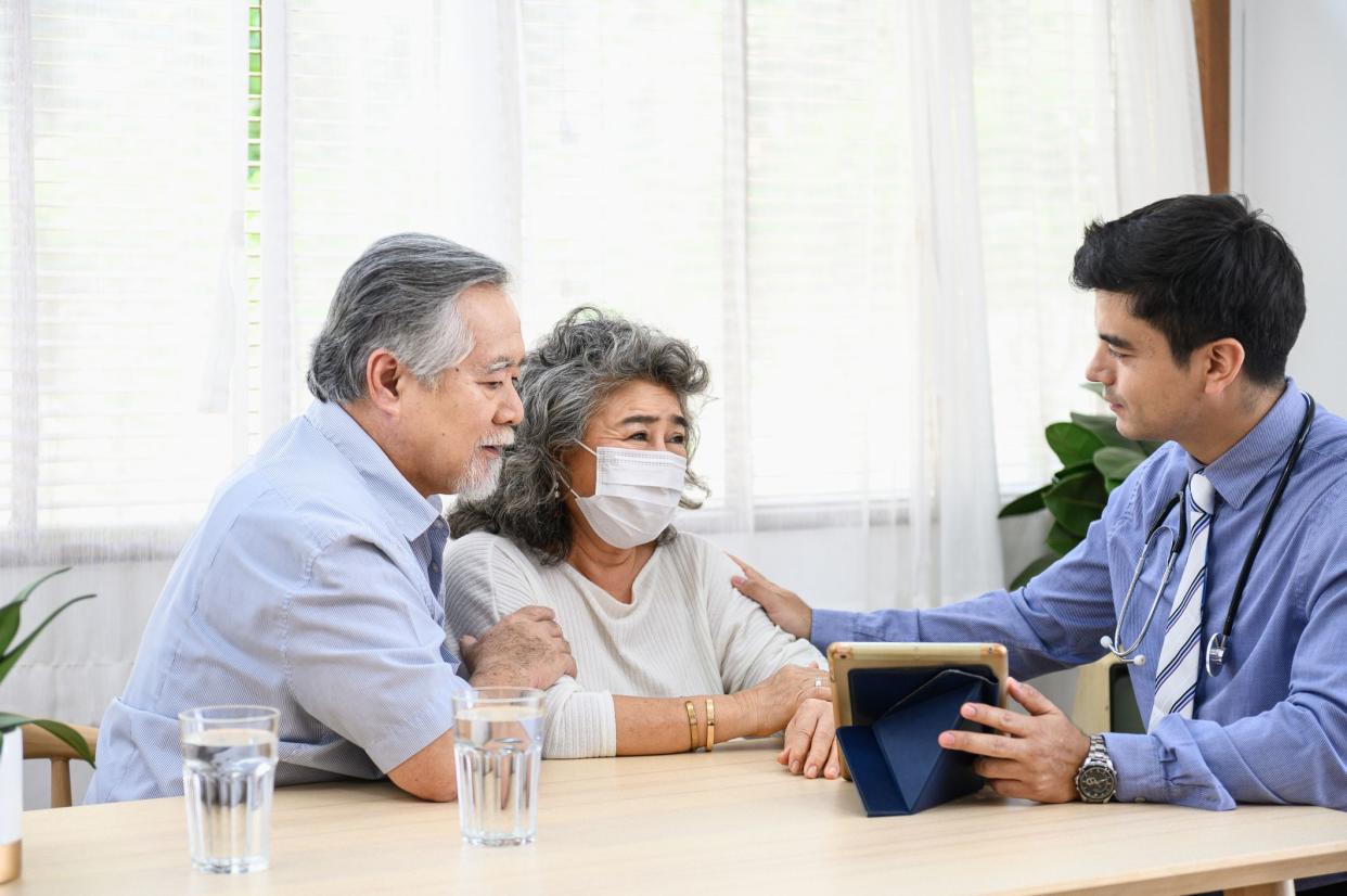 Asian senior retired couple receiving health advisory from doctor by explaining illness symptom and wearing face mask when consulting at home.