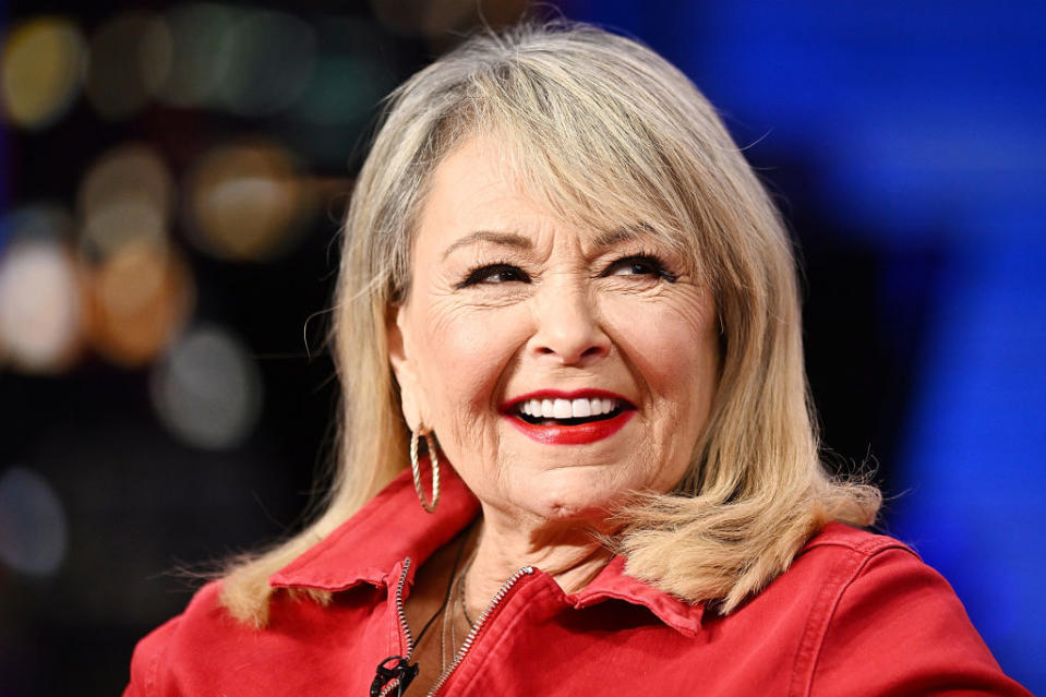 Roseanne Barr lächelt, während sie in einem Innenbereich eine lässige Jacke und Creolen trägt