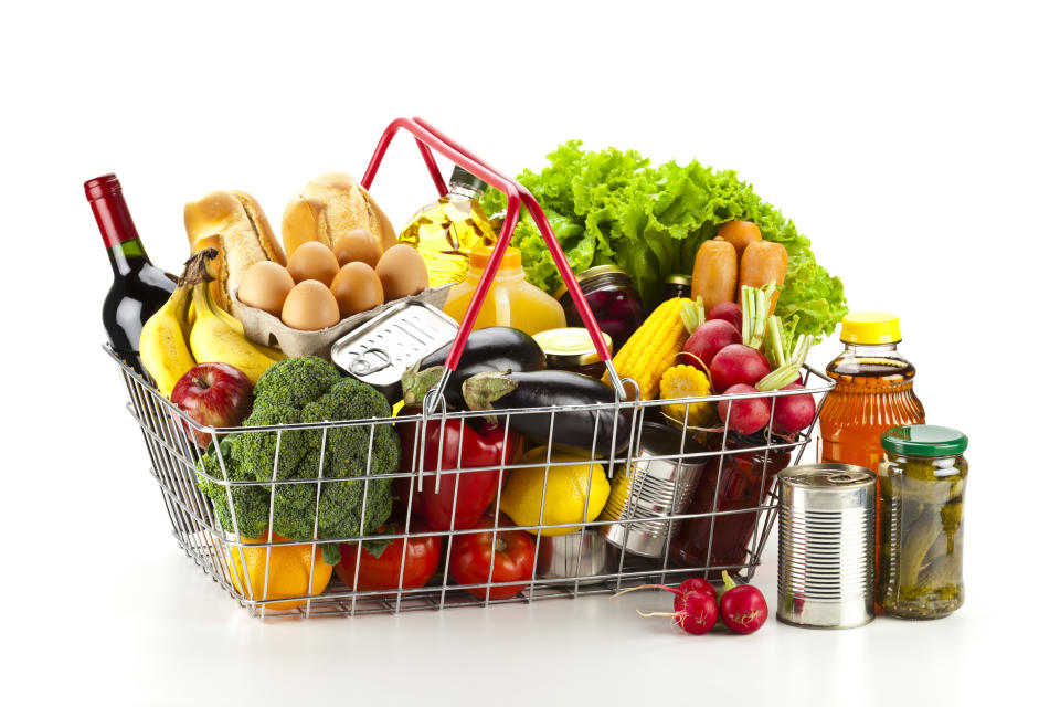 Side view of a wire shopping basket filled with all sort of food including fresh vegetables, fruits, canned food, fruit juice and dairy products.  Some canned food is outside the basket at the right side. Colors are very vibrant and saturated with greens, yellows and reds as predominant colors. The basket is isolated on white background and produces a soft shadow. Studio shot taken with DSRL Canon EOS 5D Mk II