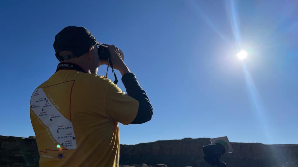 A man wearing a yellow t-shirt holds a pair of binoculars and looks at the sun.