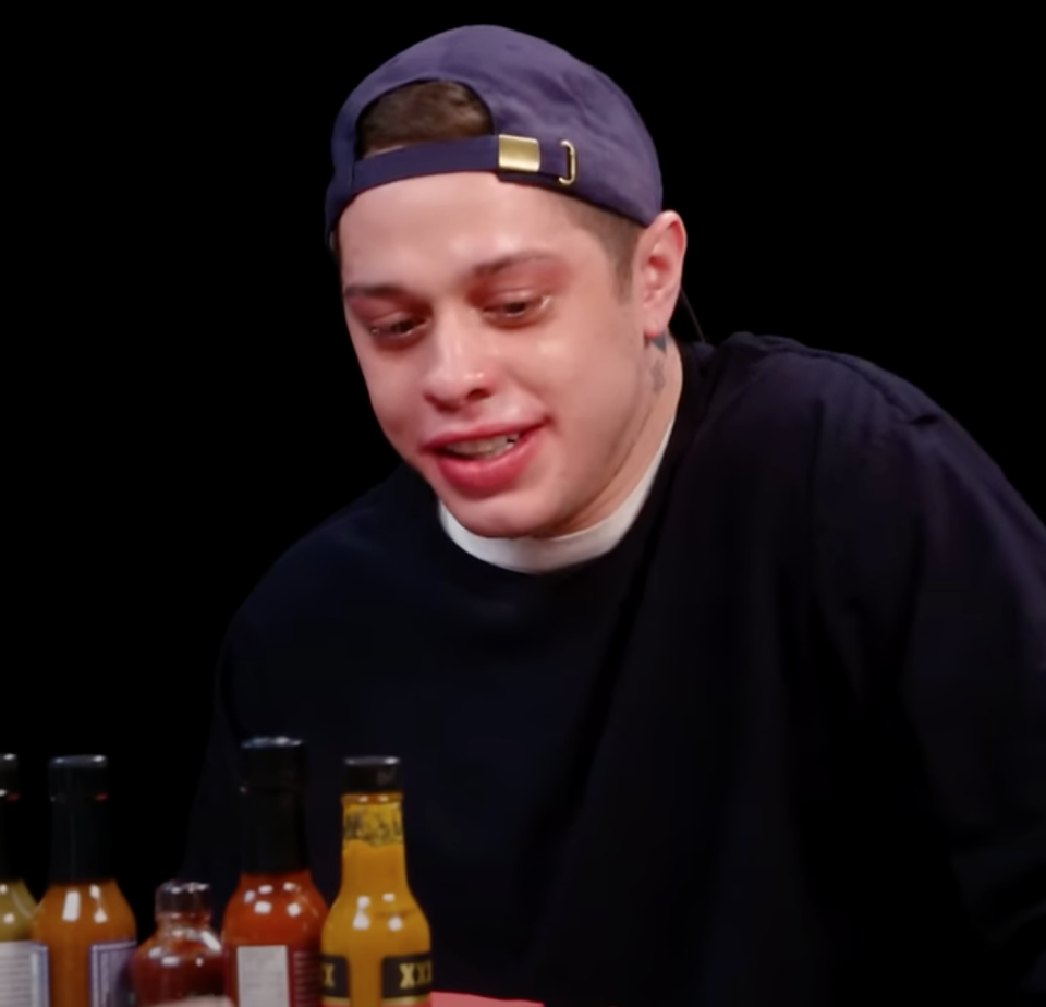 Pete Davidson seated at a table with various hot sauce bottles and a sweaty face