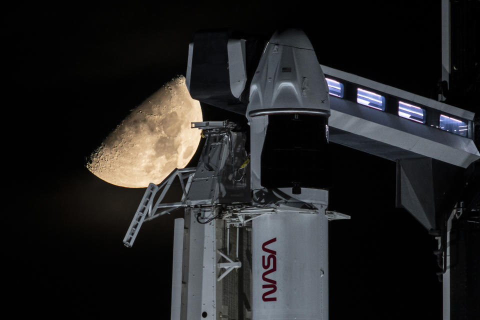 A half moon hangs behind the top of a white rocket and crew access arm