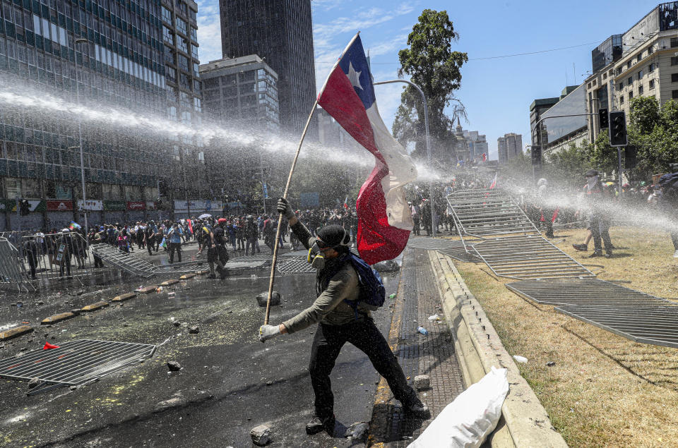 An anti-government demonstrator holds a Chilean flag as he braces himself in front of a police water cannon outside La Moneda presidential palace in Santiago, Chile, Tuesday, Nov. 12, 2019. Chile is entering its 26th day of protest against social inequality with huge demonstrations across the country and a national strike. (AP Photo/Esteban Felix)