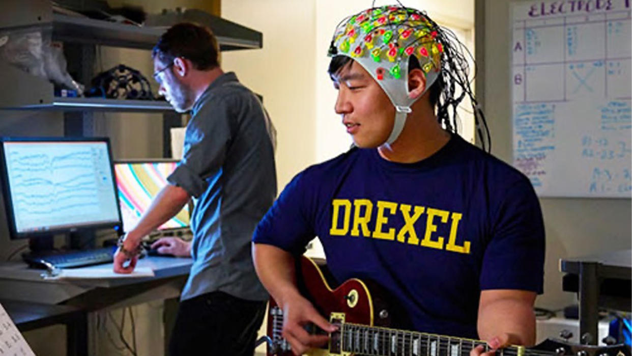  A man with tan skin and black hair wearing an EEG cap with colorful electrodes on it as he plays a guitar. A researcher stands in the background at a computer, watching brain waves on the screen. 