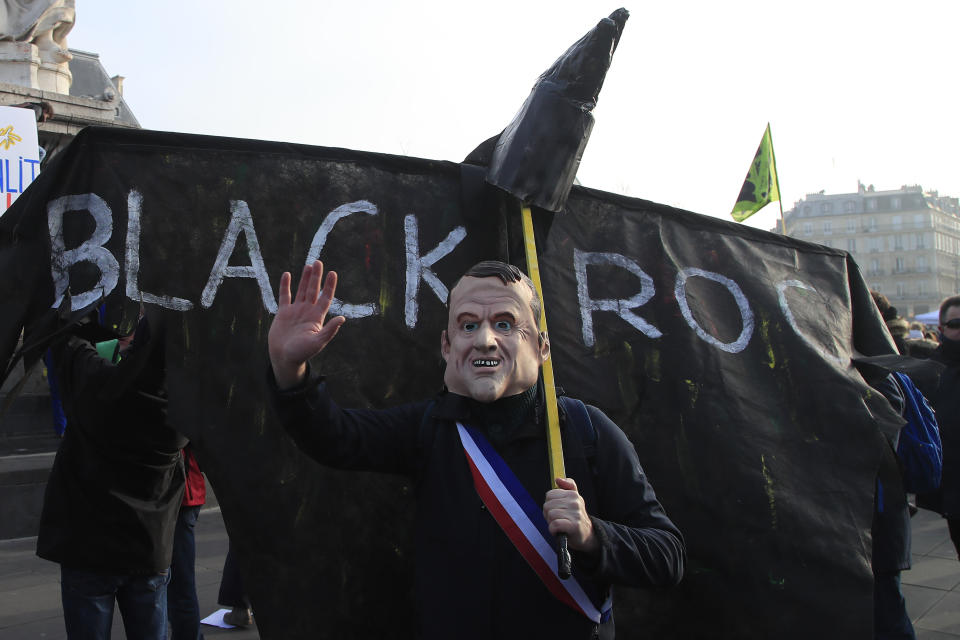 A protester wearing a mask of French President Emmanuel Macron gestures in front of a banner against US financial company BlackRock, Friday, Jan. 24, 2020 in Paris. French unions are holding last-ditch strikes and protests around the country Friday as the government unveils a divisive bill redesigning the national retirement system. (AP Photo/Michel Euler)