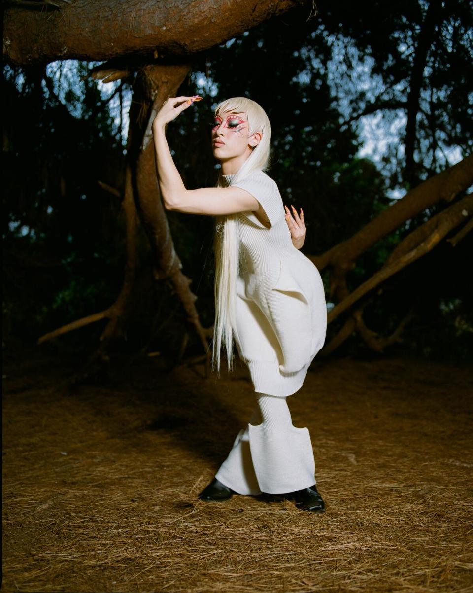 A person with long white-blond hair strikes a pose outside in a white Issey Miyake outfit.