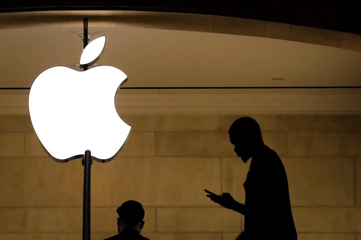 The sihlouette of a person looking at their phone while walking near an illuminated logo for Apple.