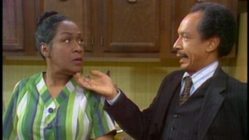 a photo of george jefferson wearing a brown suit standing beside weezy jefferson with a green dress