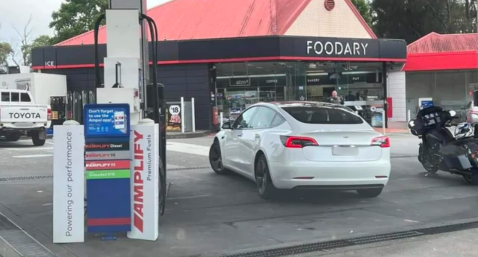 Image of a white Tesla electric vehicle parked at a fuel pump with the 'Foodary' shop in the background.